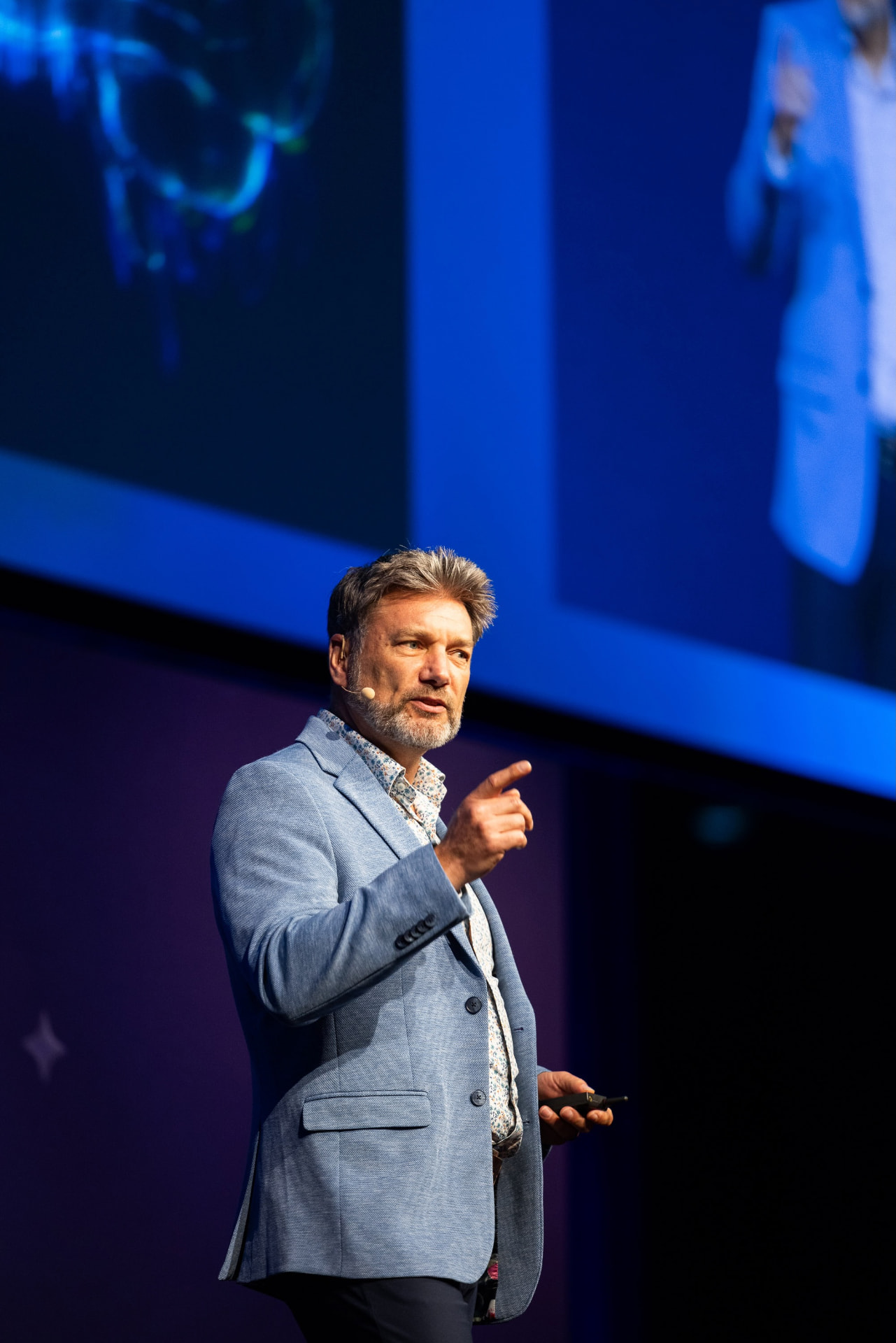 Simon Grice presenting on stage in a light blazer, gesturing toward the audience with a large blue presentation screen behind him